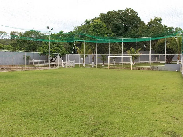Campo de futebol gramado cercado por alambrado e tela verde, com traves brancas e árvores ao fundo
